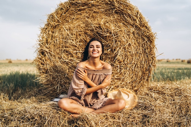 A young woman with long hair and in a dress sits near a hay bale. Woman posing smiling and looking at camera