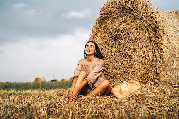 A young woman with long hair and in a dress sits near a hay bale. Woman posing smiling and looking