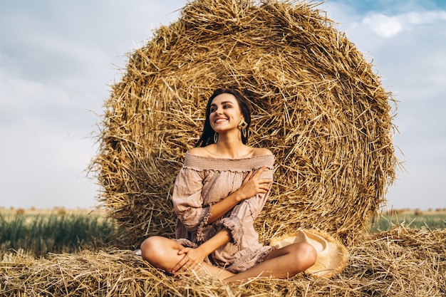 A young woman with long hair and in a dress sits near a hay bale. Woman posing smiling