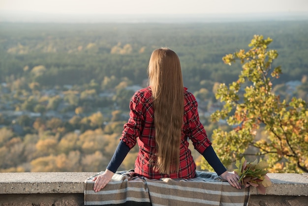 Young woman with long hair sits on a hill overlooking the village Back view