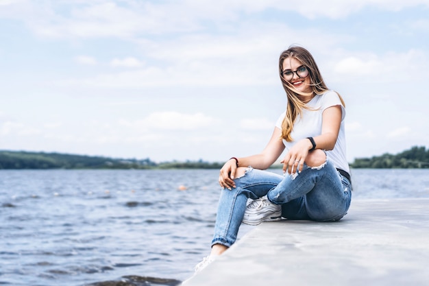 Young woman with long hair in stylish glasses posing on the concrete shore near the lake