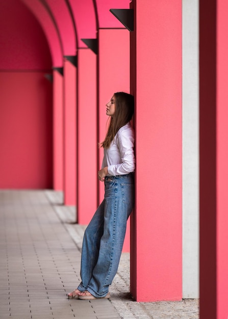 A young woman with long hair in a white blouse and blue jeans stands among pink columns looks into the distance Full length photo