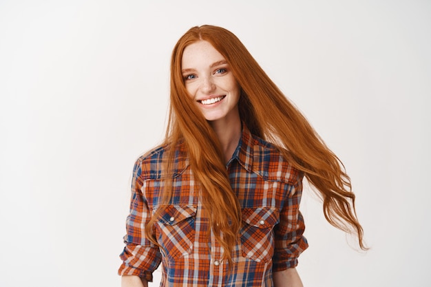 Young woman with long healthy ginger hair, smiling happy at at front, standing on white wall