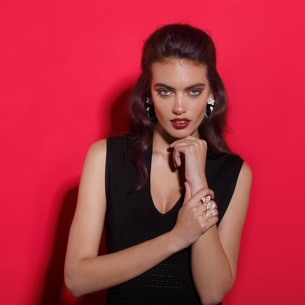 Young woman with makeup and hairstyle, red lipstick in elegant slim black dress, wears jewellery, over red background.