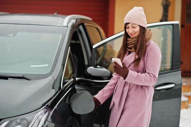 Young woman with mobile phone at hand charging her electric car in winter