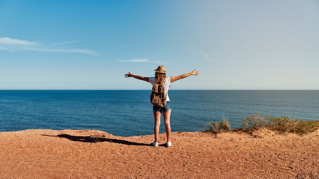 Young woman with open arms, with hat and backpack in front of the sea in summer