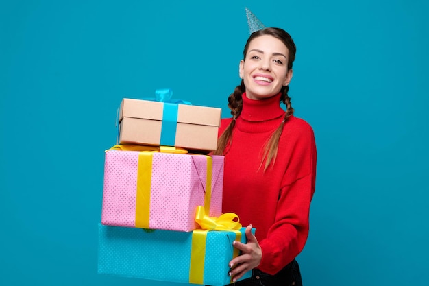 Young woman with party cone on head holding stack of gift and laughing on blue isolated background