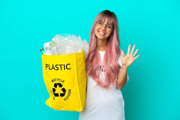 Young woman with pink hair holding a bag full of plastic bottles to recycle isolated on blue background counting five with fingers