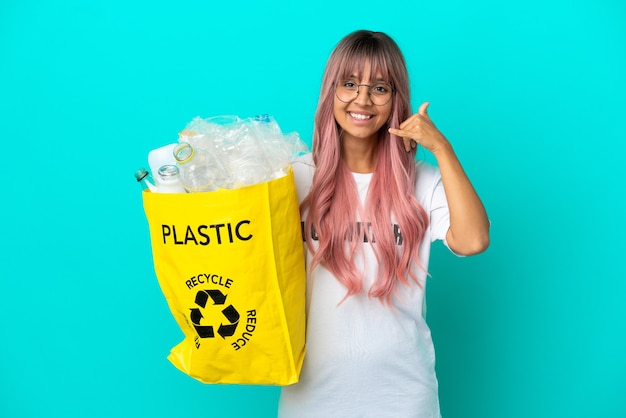 Young woman with pink hair holding a bag full of plastic bottles to recycle isolated on blue background making phone gesture. Call me back sign