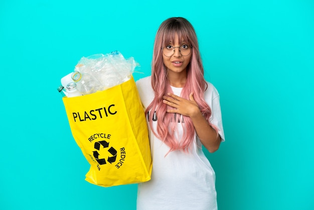 Young woman with pink hair holding a bag full of plastic bottles to recycle isolated on blue background surprised and shocked while looking right