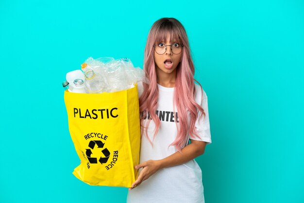 Young woman with pink hair holding a bag full of plastic bottles to recycle isolated on blue background with surprise facial expression