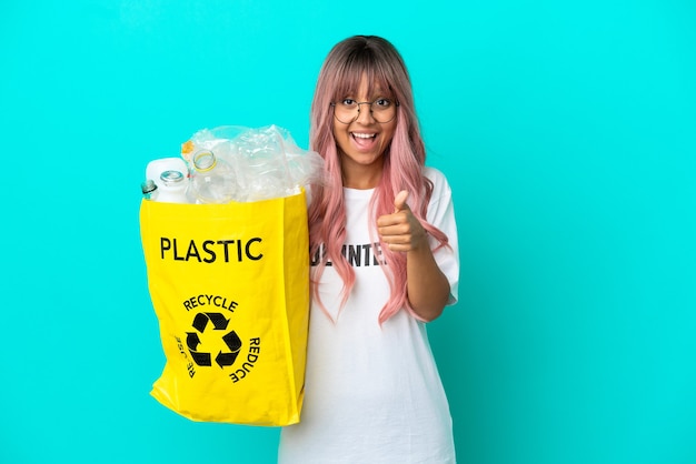 Young woman with pink hair holding a bag full of plastic bottles to recycle isolated on blue background with thumbs up because something good has happened