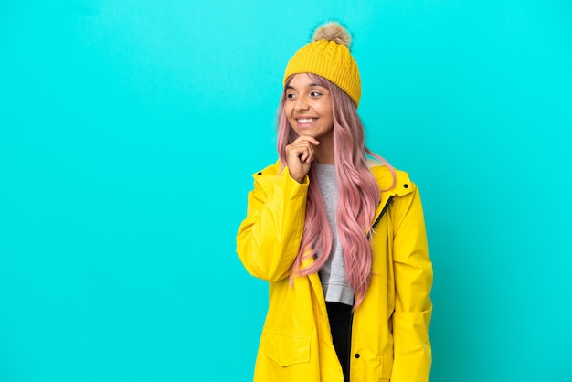 Young woman with pink hair wearing a rainproof coat isolated on blue background looking to the side and smiling