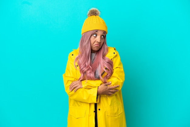 Young woman with pink hair wearing a rainproof coat isolated on blue background making doubts gesture while lifting the shoulders