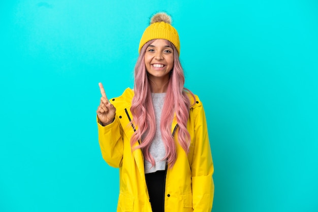 Young woman with pink hair wearing a rainproof coat isolated on blue background pointing up a great idea