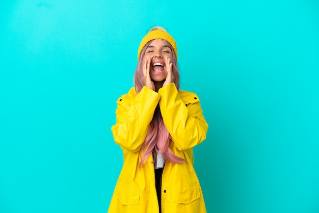 Young woman with pink hair wearing a rainproof coat isolated on blue background shouting and announcing something