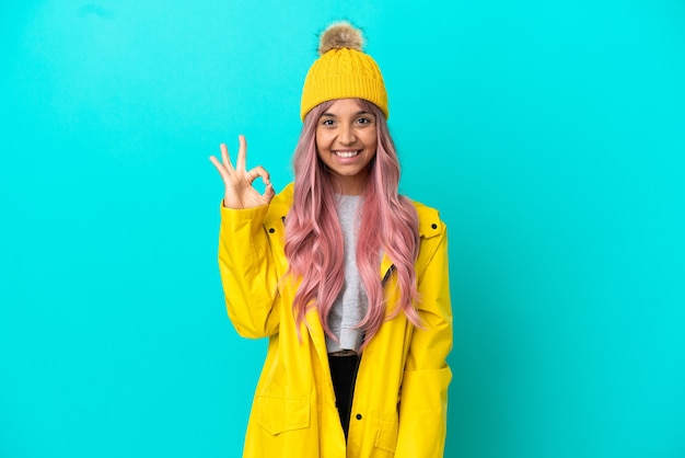 Young woman with pink hair wearing a rainproof coat isolated on blue background showing ok sign with fingers