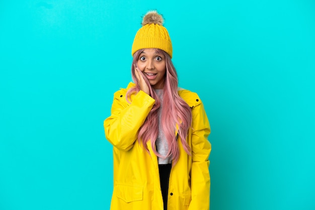 Young woman with pink hair wearing a rainproof coat isolated on blue background with surprise and shocked facial expression