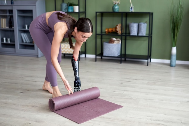 Young woman with prosthetic arm finishing her training at home rolling yoga exercise mat