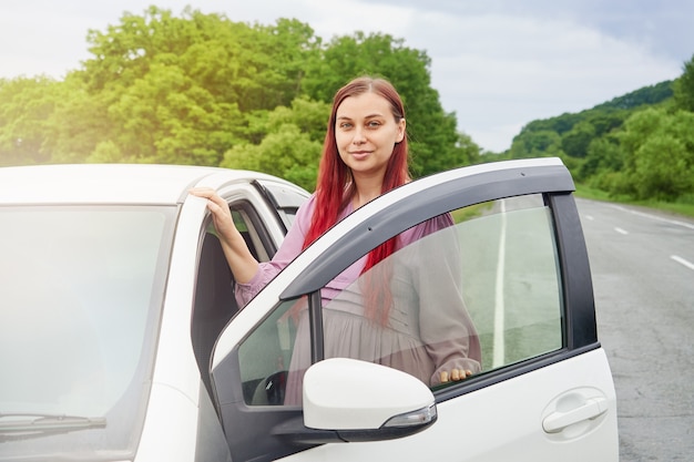 Young woman with red hair stands next to the car opening the door, parked on the side of the road.