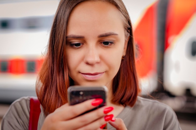 Young woman with short hair stands on platform browsing mobile phone waiting for train Portrait of female passenger using smartphone on background of blurry train