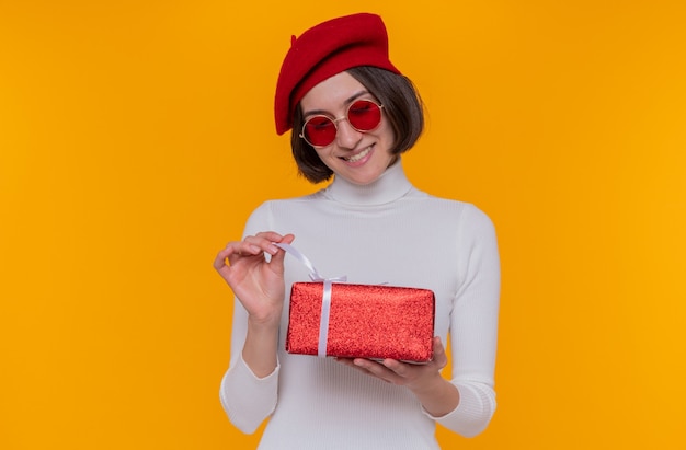 young woman with short hair in white turtleneck wearing beret and red sunglasses holding a present happy and cheerful going to open present smiling cheerfully