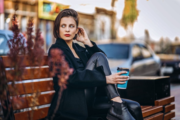 Young woman with short haircut and red lips, dressed in black coat, sits on a bench in the city and holds a cup of coffee.