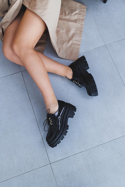 A young woman with slender beautiful legs in fashionable black leather shoes sits on a chair in the studio Womens stylish loafers closeup Spring shoe collection