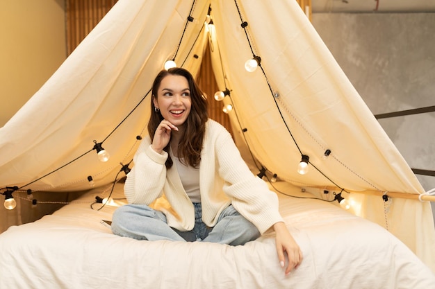 A young woman with a slight smile on her face sits on a canopy bed with cozy light bulbs