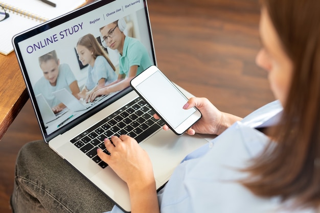 Young woman with smartphone sitting in front of laptop display while surfing on educational website