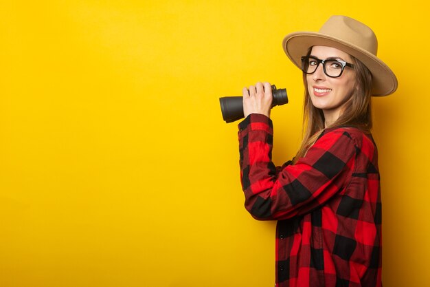 Young woman with a smile in a hat and a plaid shirt holding binoculars in her hands on a yellow surface