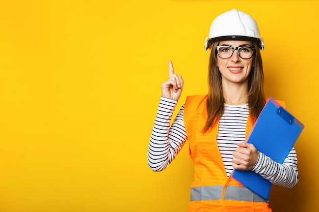 Young woman with a smile in a vest and hard hat holds a clipboard on a yellow background Construction concept new building Banner