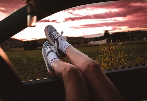 Young woman with sneakers with feet propped on the car window at sunset