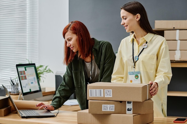 Young woman with stack of packed boxes standing by colleague with laptop