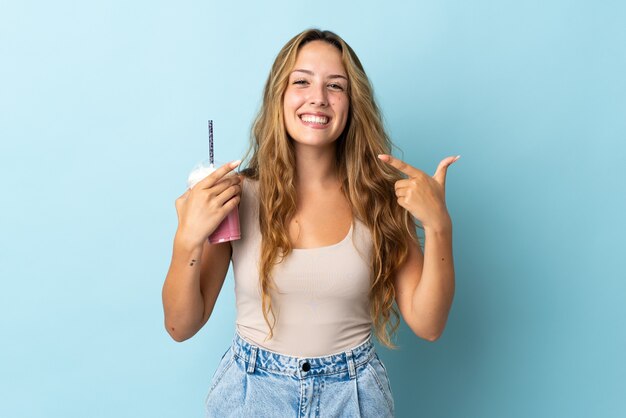 Young woman with strawberry milkshake isolated on blue wall giving a thumbs up gesture