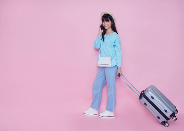 Young woman with suitcase and talking on phone on pink wall