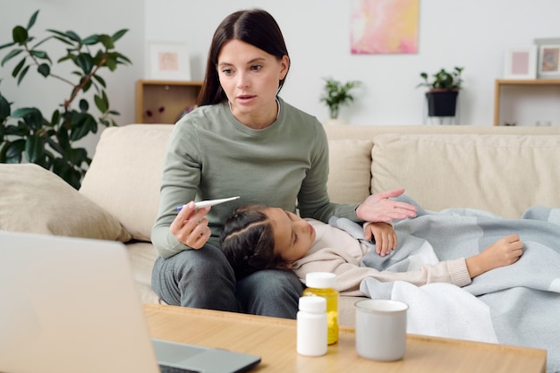 Young woman with thermometer describing symptoms of sick little daughter to online doctor while sitting on couch next to her