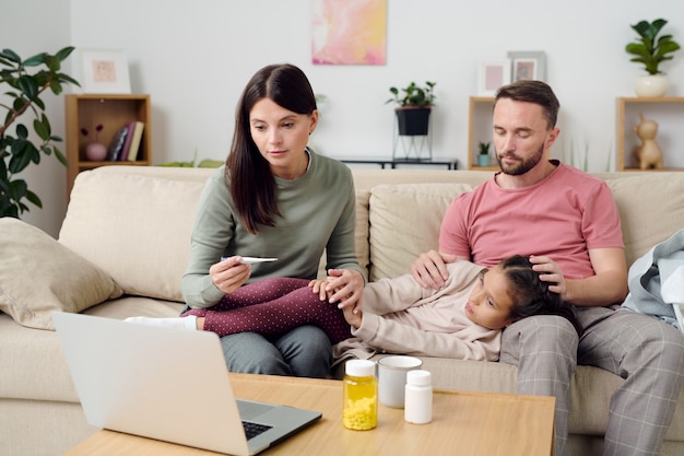 Young woman with thermometer describing symptoms of sick little daughter to online doctor while sitting next to her and husband