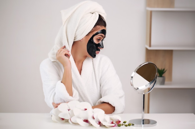 Young woman with towel on head and black face mask sitting at home