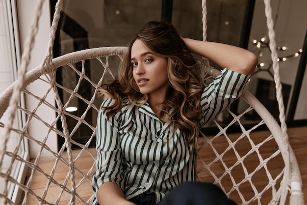 Young woman with wavy hair looks into camera in apartment Tanned Caucasian girl in silk pajamas si