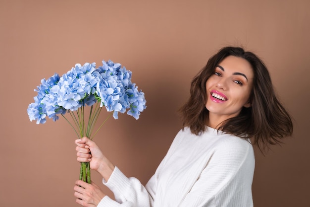 A young woman with wavy voluminous hair on a beige background with bright pink lipstick lip gloss in a white sweater holds a bouquet of blue flowers