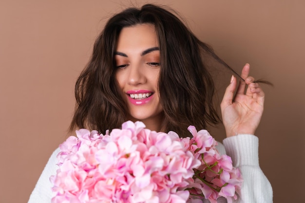 A young woman with wavy voluminous hair on a beige background with bright pink lipstick lip gloss in a white sweater holds a bouquet of pink flowers