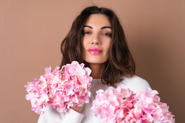 A young woman with wavy voluminous hair on a beige background with bright pink lipstick lip gloss in a white sweater holds a bouquet of pink flowers