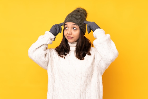 Young woman with winter hat over isolated yellow wall having doubts and thinking