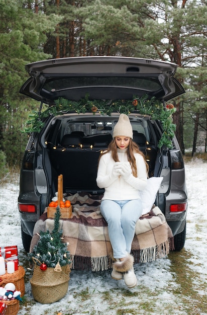 A young woman in a wool hat sits in the trunk of a car and holds a Cup of hot tea