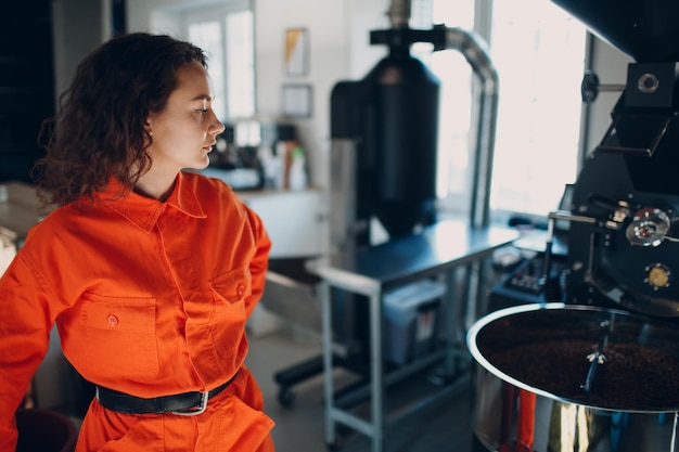 Young woman worker in orange suit in workshop with Coffee roaster machine during coffee roasting process