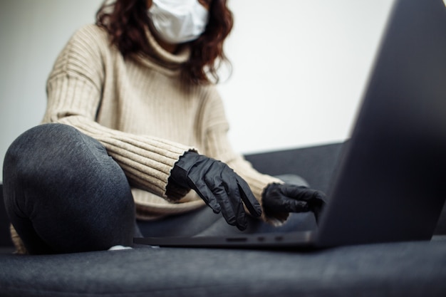 Young woman working from home during quarantine due to coronavirus pandemia. Beautiful girl stays home wearing medical mask and gloves and typing on a laptop. Covid-19 epidemia worldwide concept.