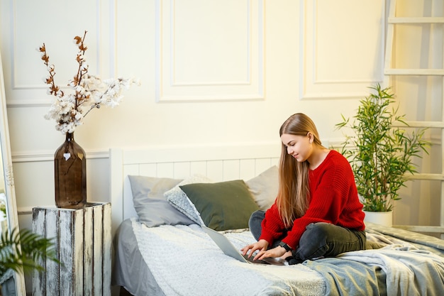 Young woman works at a computer, sitting on a bed, working from a distance. A girl with long hair in a red sweater and jeans works at home.