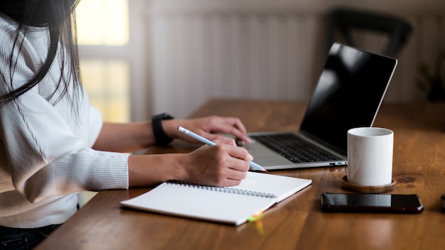 Young woman works from home, she uses a laptop and takes notes, a coffee mug and a smartphone on the table.