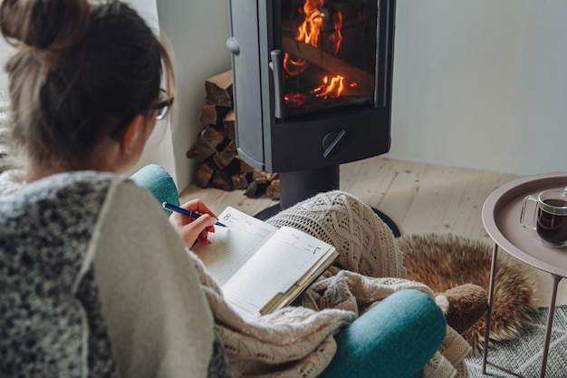 Young woman write in notebook sitting in armchair by fireplace